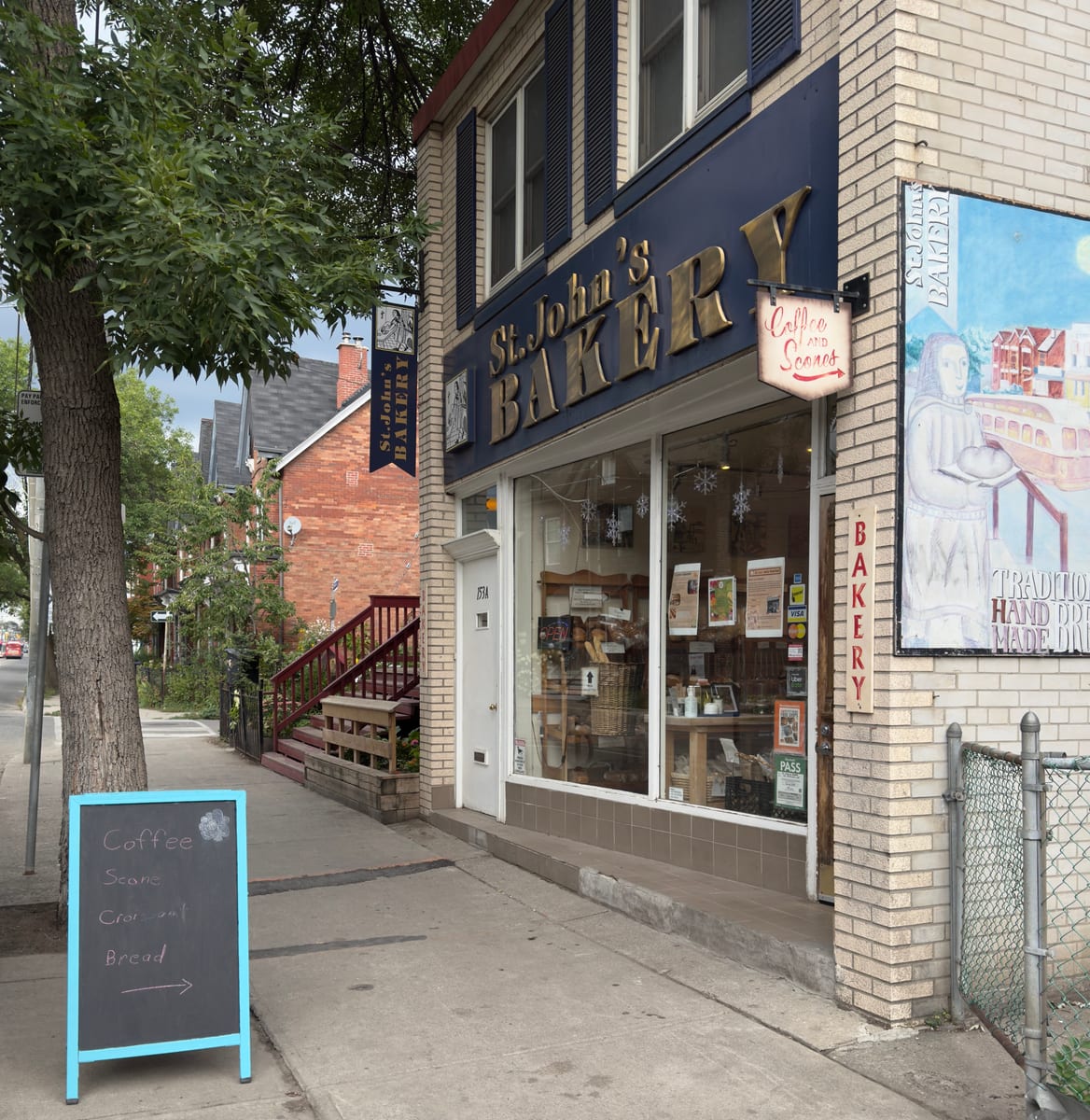 The St. John's Bakery storefront on Broadview Avenue, gold letters on a navy fascia, a mural on the side wall and a chalkboard sign on the sidewalk