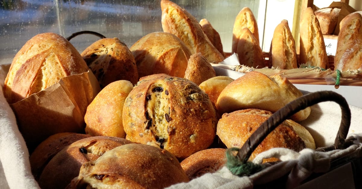 A cloth-lined basket of crusty rolls and loaves in low sun by the shop window, more loaves standing behind it