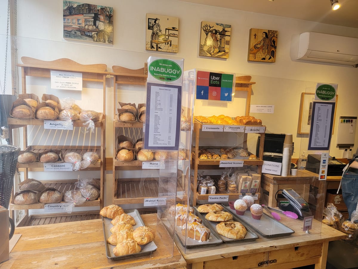 The shop counter at St. John's Bakery: wooden shelves of labelled sourdough loaves, trays of croissants, scones and cakes, and a price list on the counter