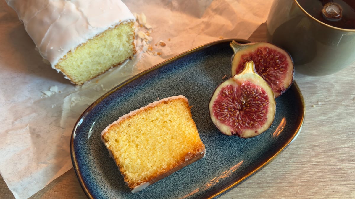 A slice of glazed pound cake on a blue plate beside a halved fig, the rest of the loaf on paper behind it